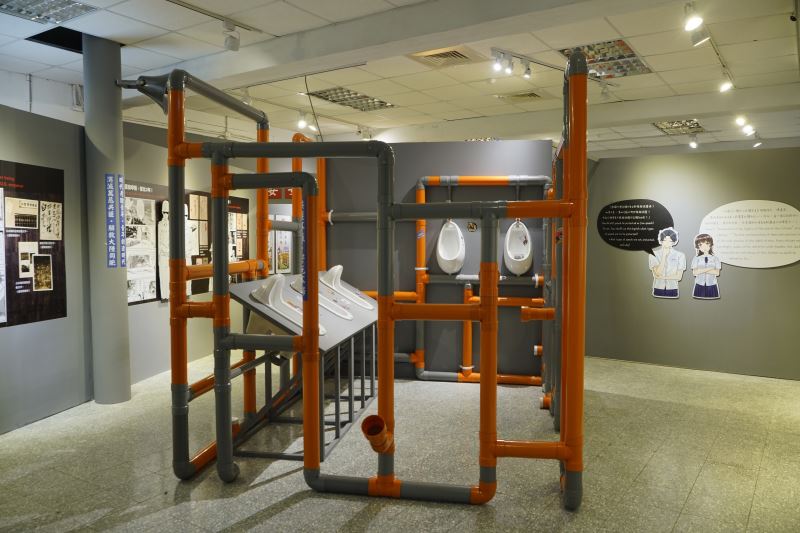 藝術家蔡坤霖搭建一座流竄著不能說的秘密的公廁空間,藉由污水管傳出的各種聲音,體驗平行時空隔牆有耳的荒謬錯亂